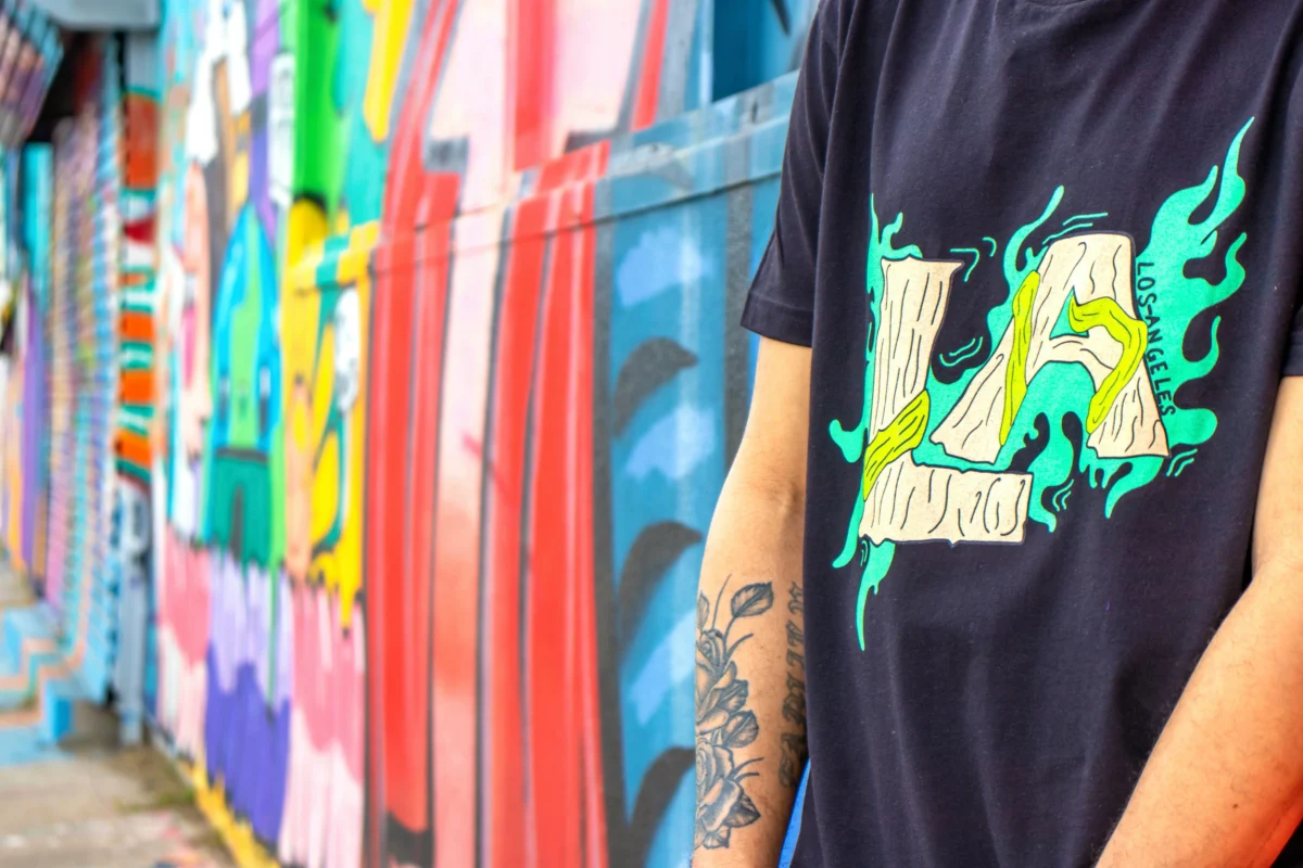 A young man wearing a graphic tee with jeans and sneakers, standing against a graffiti wall.